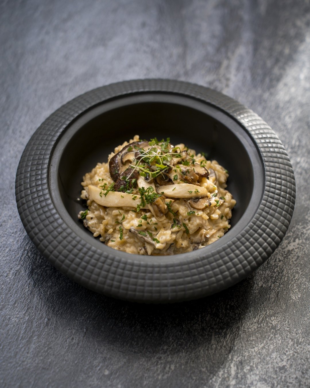 A bowl of creamy mushroom risotto garnished with chopped herbs in a black textured bowl on a dark surface.