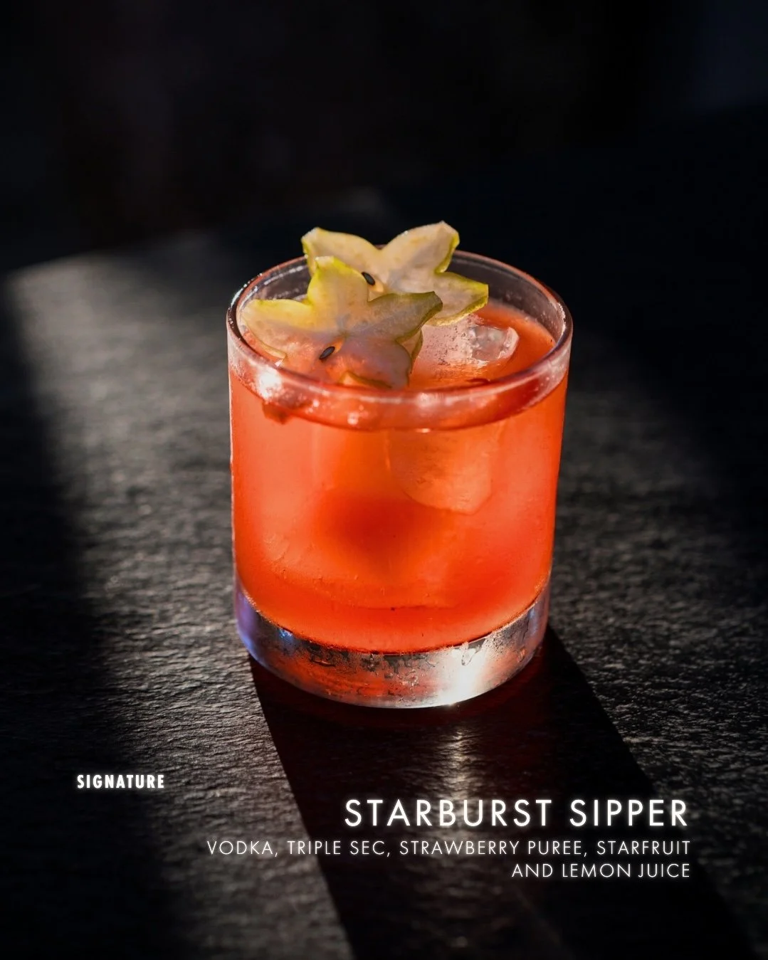 A pinkish cocktail in a short glass with starfruit slices on top, ice cubes, and condensation on the outside of the glass, set on a dark surface.