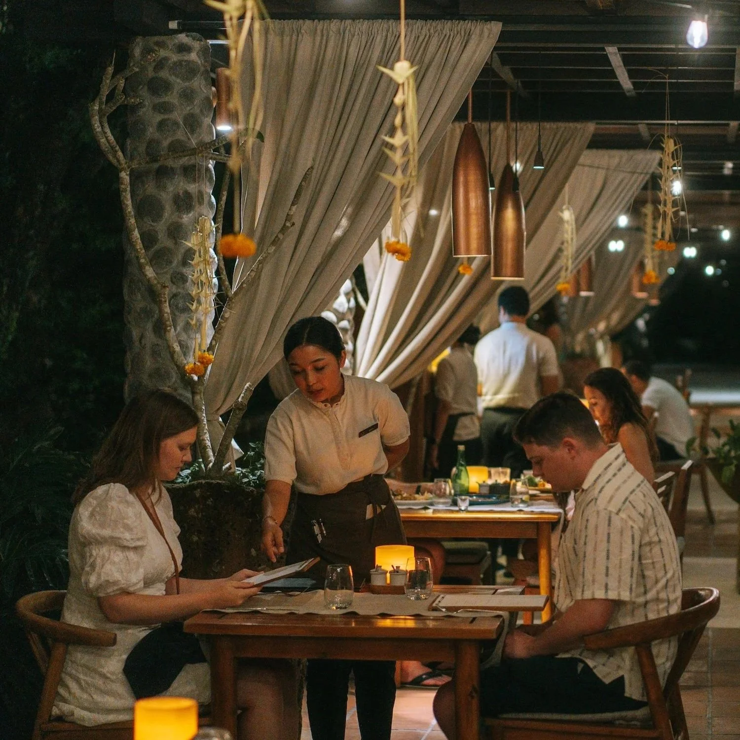 A couple dining at one of the best restaurants in Ubud at night, with a waitress taking their order, and other diners visible in the background, surrounded by warm lighting and elegant decor.