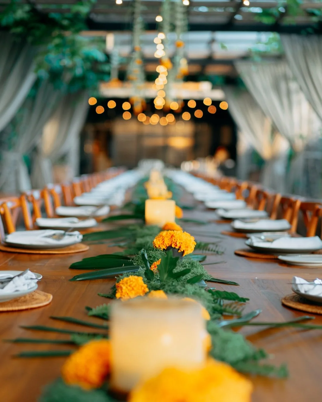 A long wooden dining table decorated with yellow marigold flowers, candles, and green foliage, set for a meal in an outdoor or semi-outdoor space with draped curtains and hanging string lights at Ubud's best restaurant.