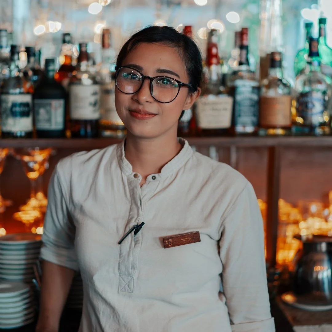 A woman with glasses and a beige uniform standing behind a bar or restaurant counter with bottles and glasses in the background.