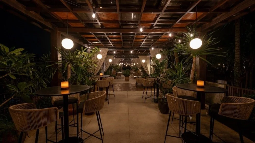 Outdoor restaurant or lounge area at night with round tables, chairs, potted plants, string lights, and candles.