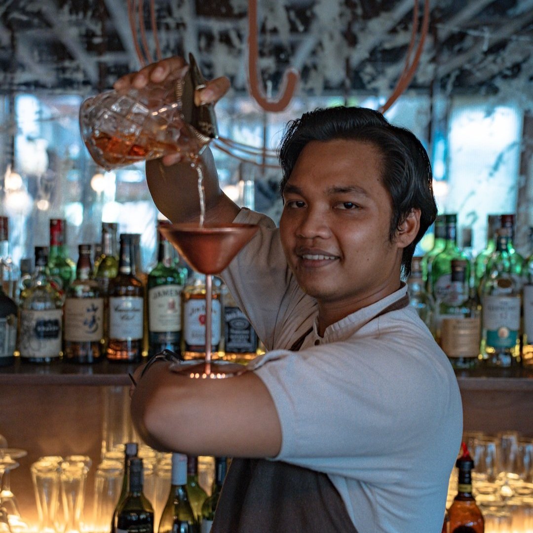 Bartender pouring a pink cocktail through a strainer at a bar.