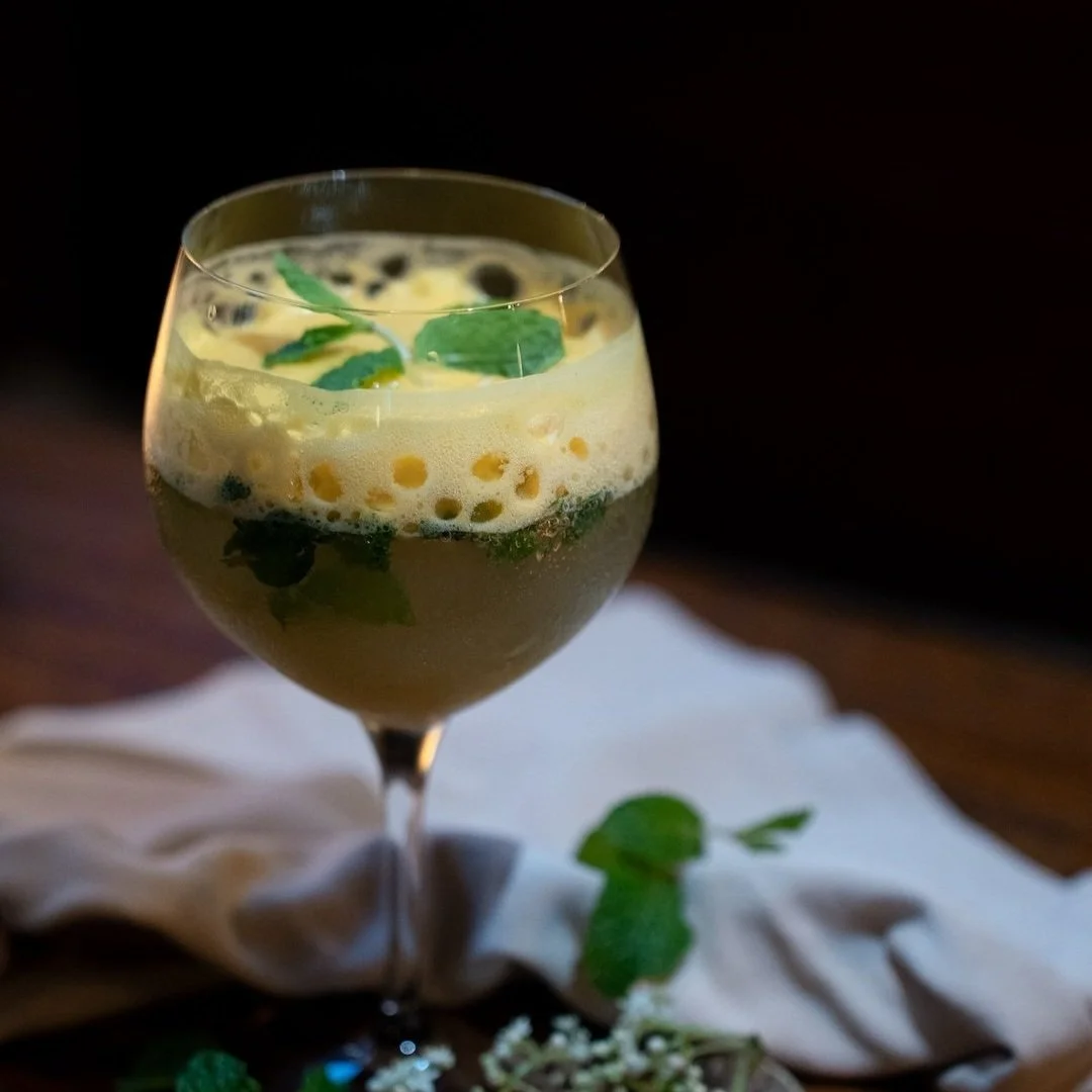 Glass of a yellow-green cocktail with mint leaves and black seeds, on a white cloth with additional mint leaves and small flowers around it.