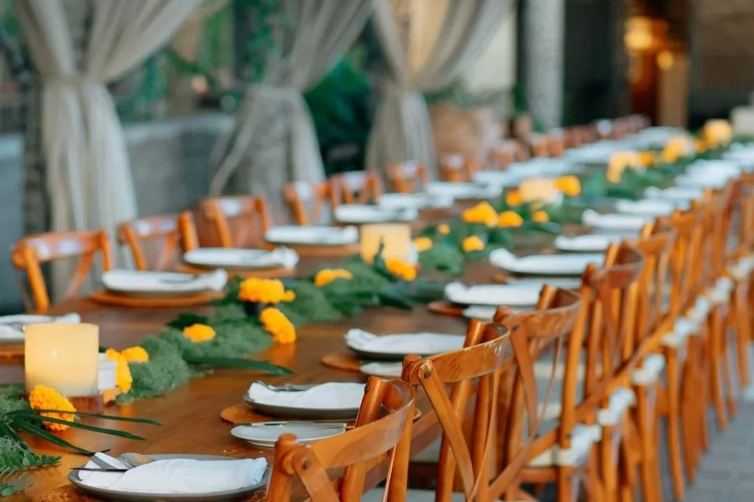 Long wooden group dining table set for a gathering with white plates, folded napkins, and green foliage with yellow flowers as decoration.