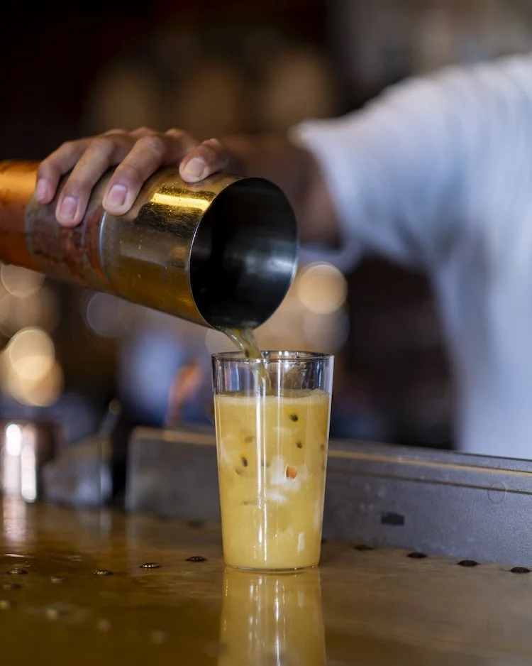 A bartender pouring passion fruit juice into a tall glass with passion fruit seeds, on a wooden bar counter.