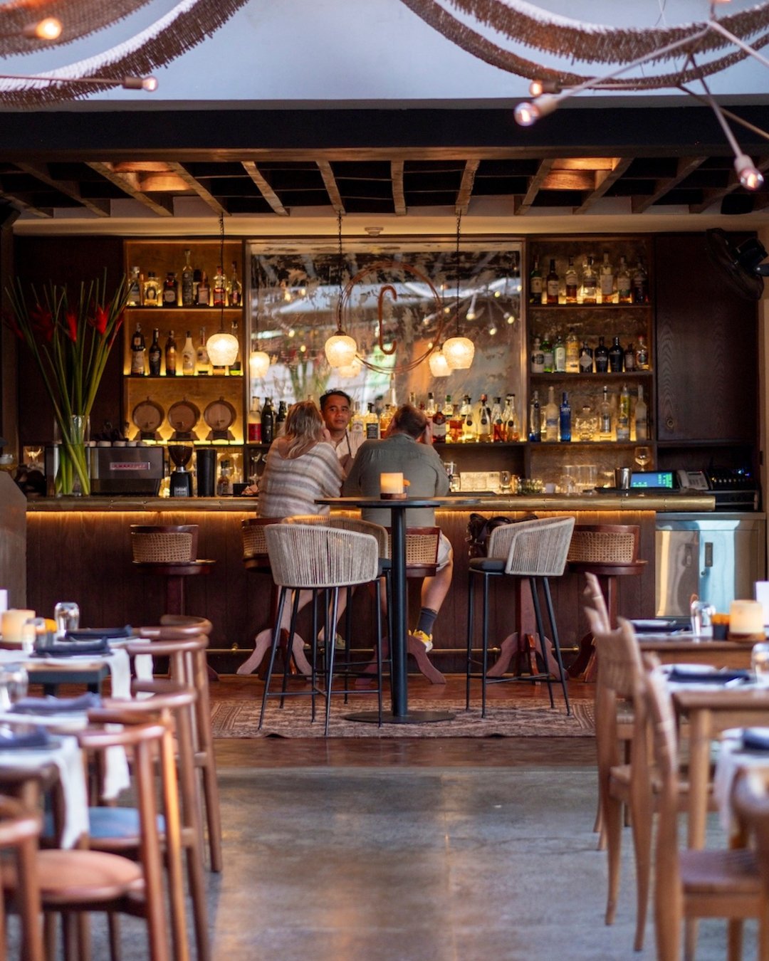 People sitting at a bar in a cozy restaurant with warm lighting, wooden furniture, and shelves of liquor bottles in the background.