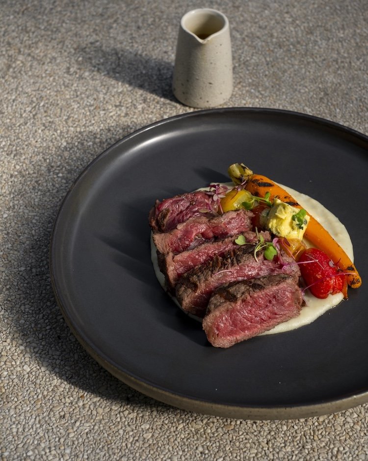 Slices of medium-rare steak served on a black plate with grilled vegetables, mashed potatoes, and microgreens, with a small ceramic pitcher in the background on a textured surface.