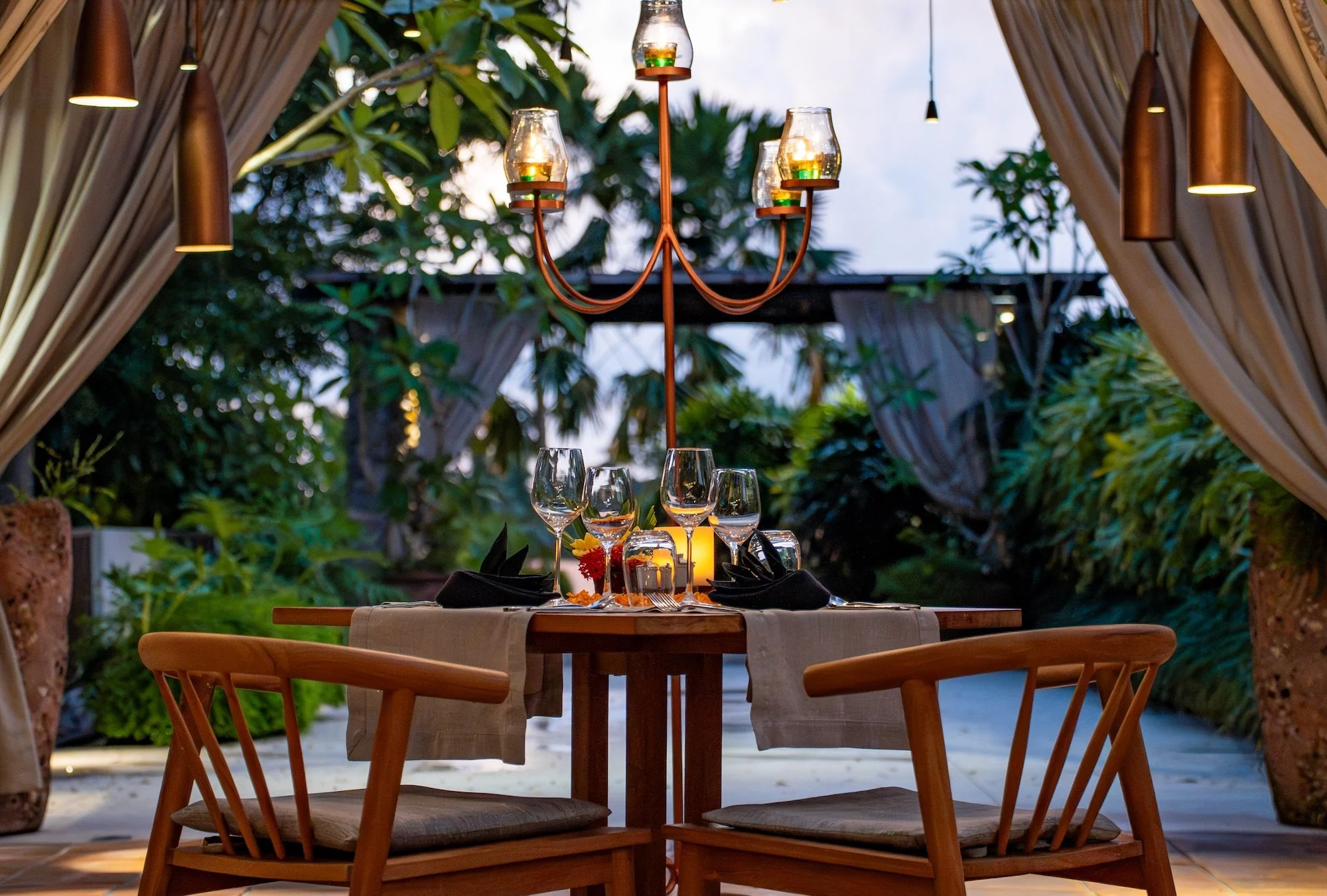 A beautifully set outdoor rooftop romantic dinner table with six wine glasses, black napkins, and a candle in the center, under a chandelier with six lit candles, surrounded by lush green tropical plants and draped curtains.