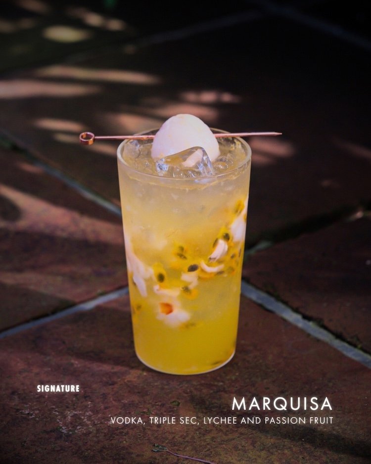 Tall glass of Marquisa cocktail with lychee and passion fruit, ice, and a lychee on a skewer, on a brick table at night.