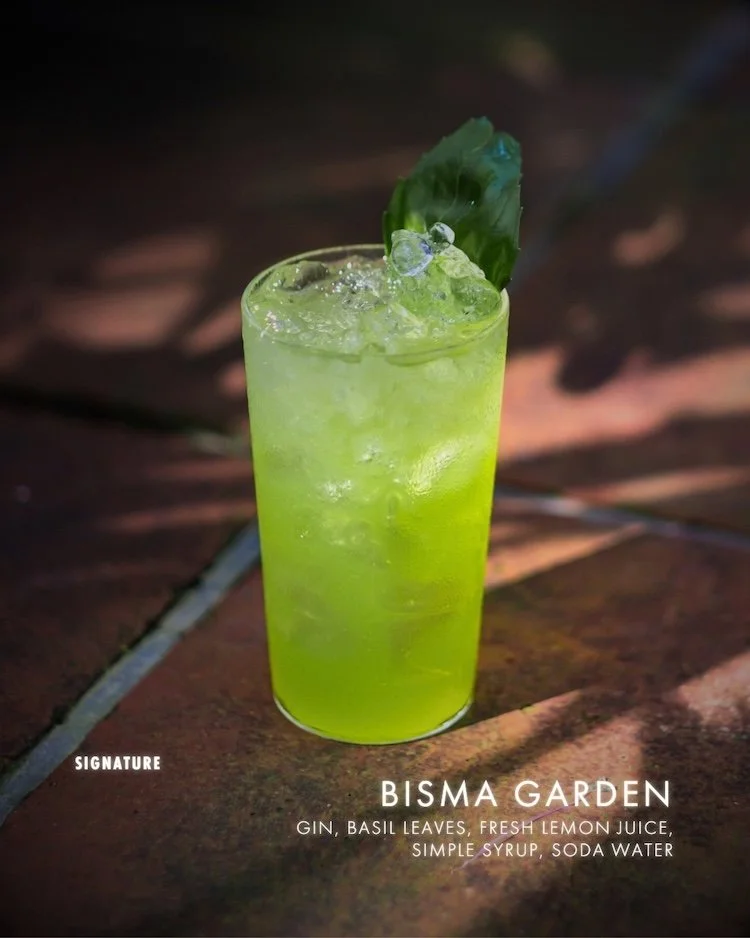 A tall glass of green cocktail garnished with a basil leaf and ice cubes, sitting on a brick surface.