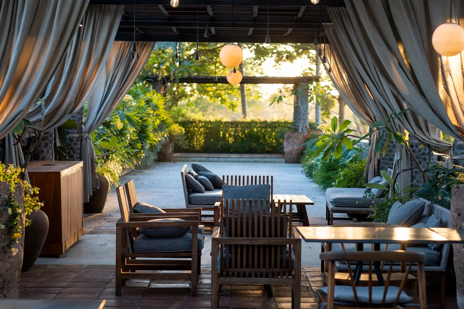 An outdoor patio with wooden chairs and sofas arranged around a coffee table, surrounded by potted plants, with beige curtains tied back on the sides, and hanging spherical lights, overlooking a lush garden at sunset.