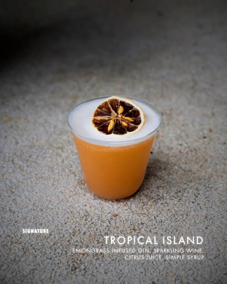 A cocktail in a clear glass with a dried citrus slice garnish on top, placed on a sandy surface.