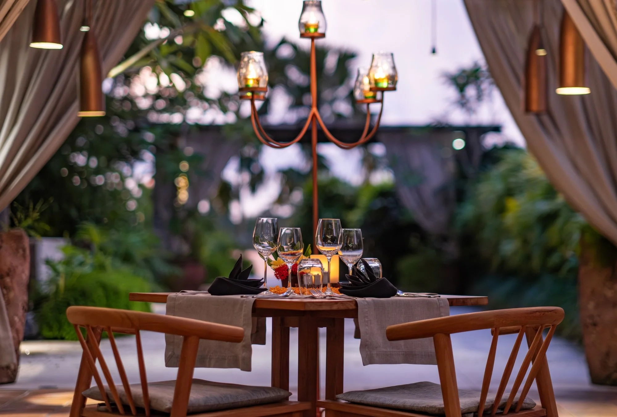 A rooftop romantic dining table set for four with wine glasses, black napkins, and a candle centerpiece, under a chandelier in an outdoor setting with lush greenery and draped curtains.
