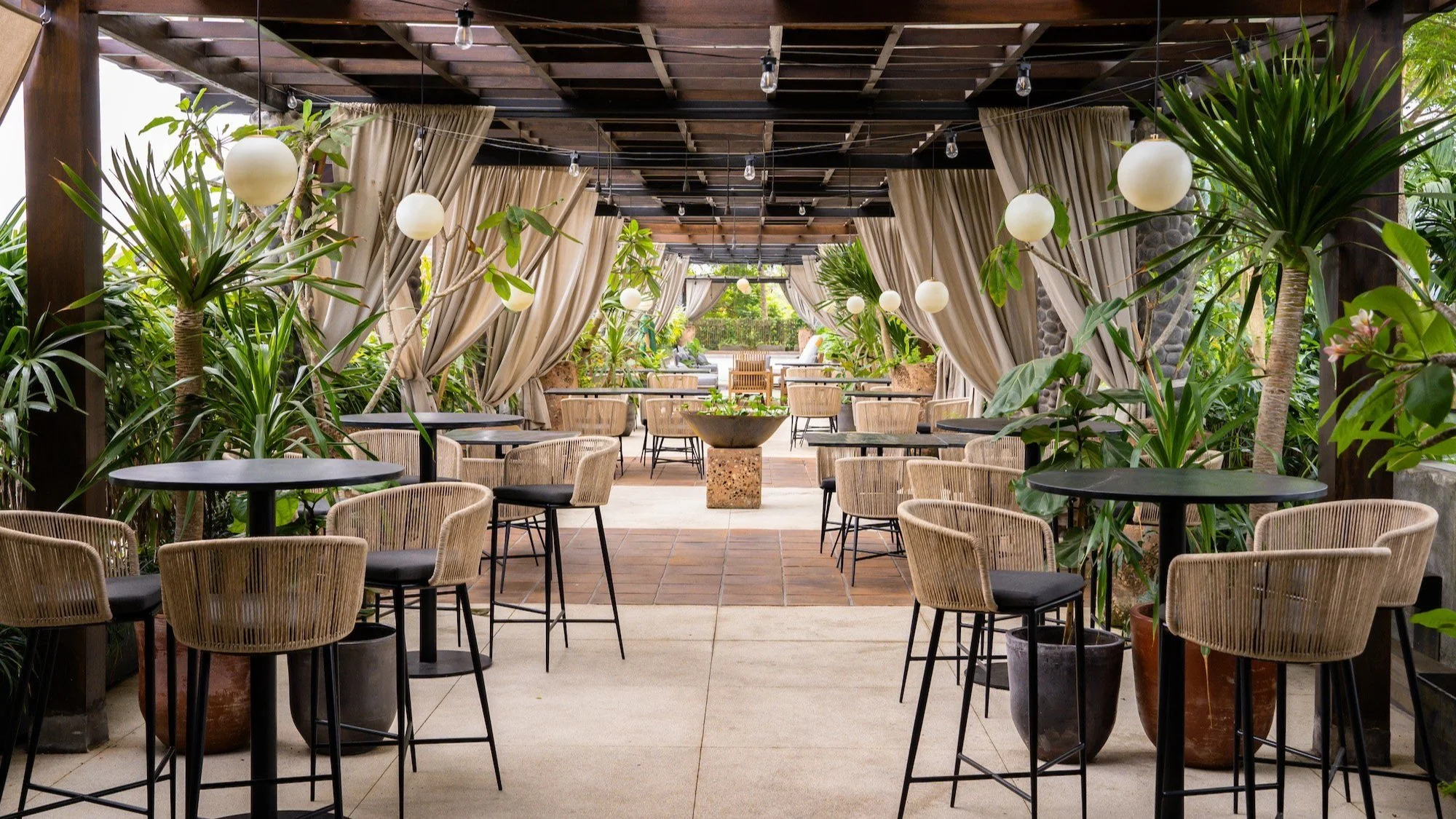An outdoor patio with round black tables, wicker chairs with black cushions, string lights, lush potted plants, and beige curtains, overlooking a garden area.