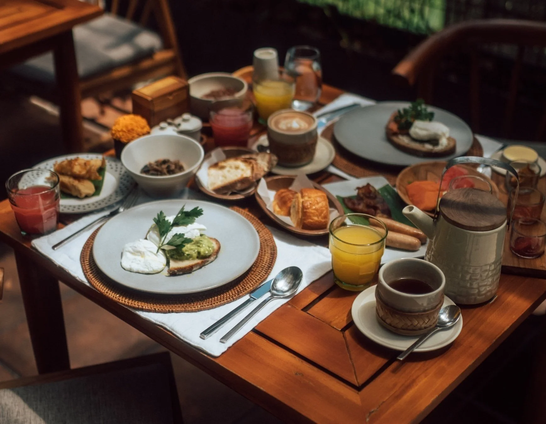 Breakfast table setting with plates of poached eggs, avocado toast, assorted pastries, cut fruits, and drinks including coffee, orange juice, and pink juice.