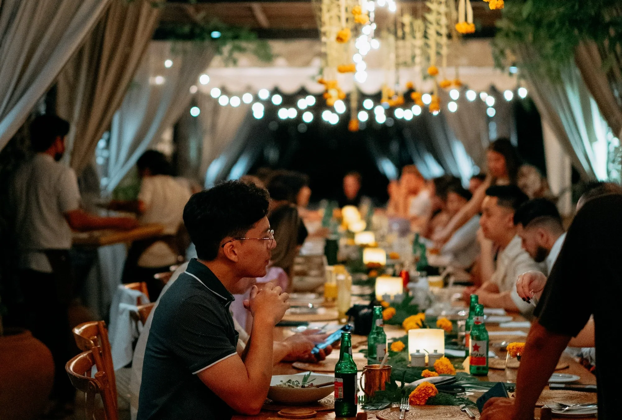 A group of people gathered around a long dinner table at night. The table is decorated with flowers, lit candles, and bottles of beverages. A festive atmosphere at Copper Ubud rooftop restaurant.