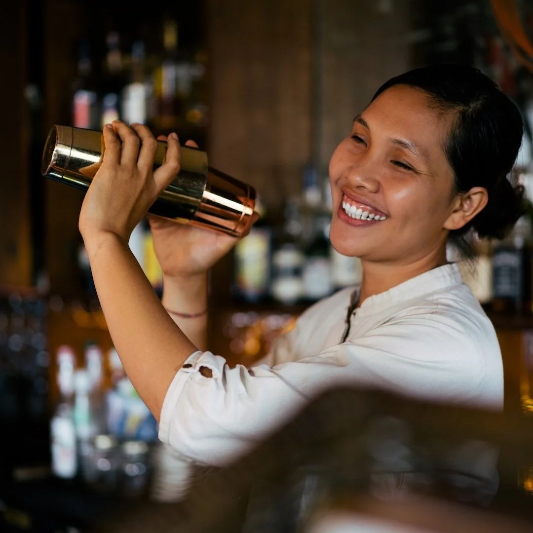 A smiling woman behind a bar, shaking a cocktail shaker.
