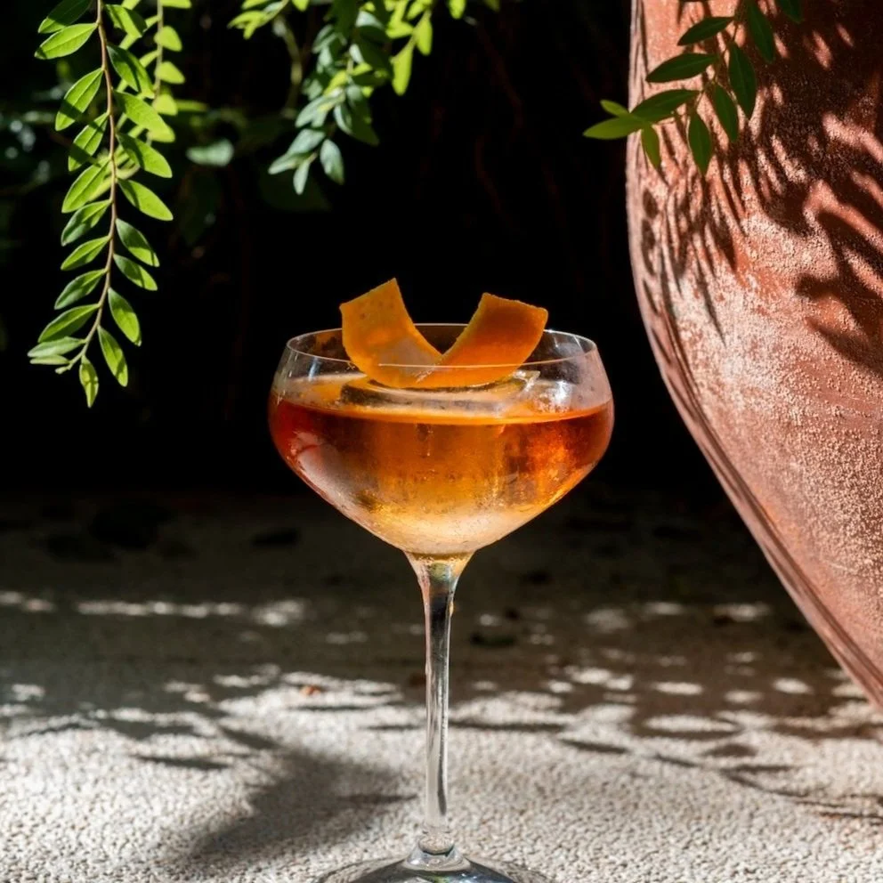 A cocktail in a coupe glass garnished with orange peel, set on a textured surface with leaf shadows, next to a large textured pot with plant leaves.
