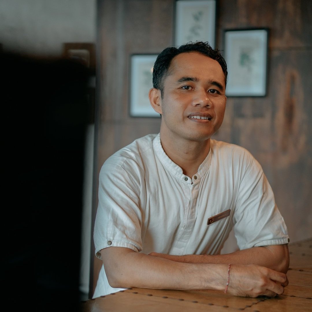 A man with short black hair wearing a cream-colored shirt sitting at a wooden table with a smile, in a room with framed pictures on the wall.