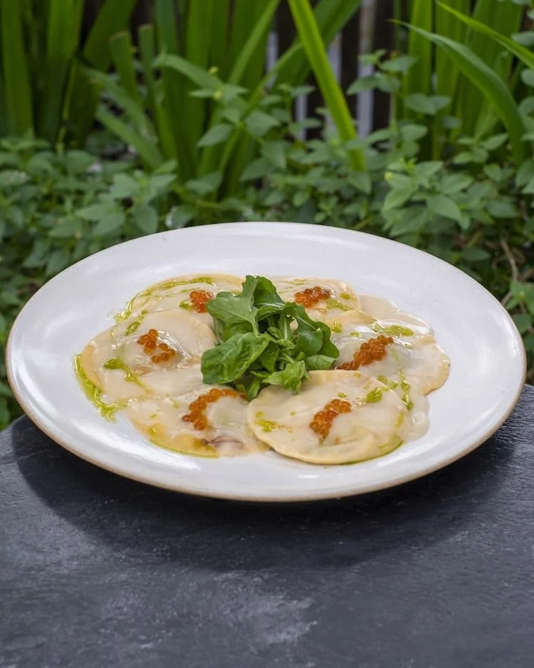 Plate of ravioli with cream sauce, garnished with green herbs and red roe, on a black surface with green plants in the background served at Copper rooftop Ubud restaurant