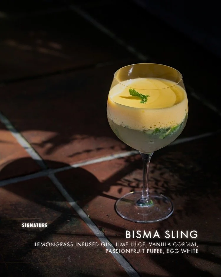 A cocktail in a stemmed glass with a lemon slice and a mint leaf, set on a tiled surface with shadows.