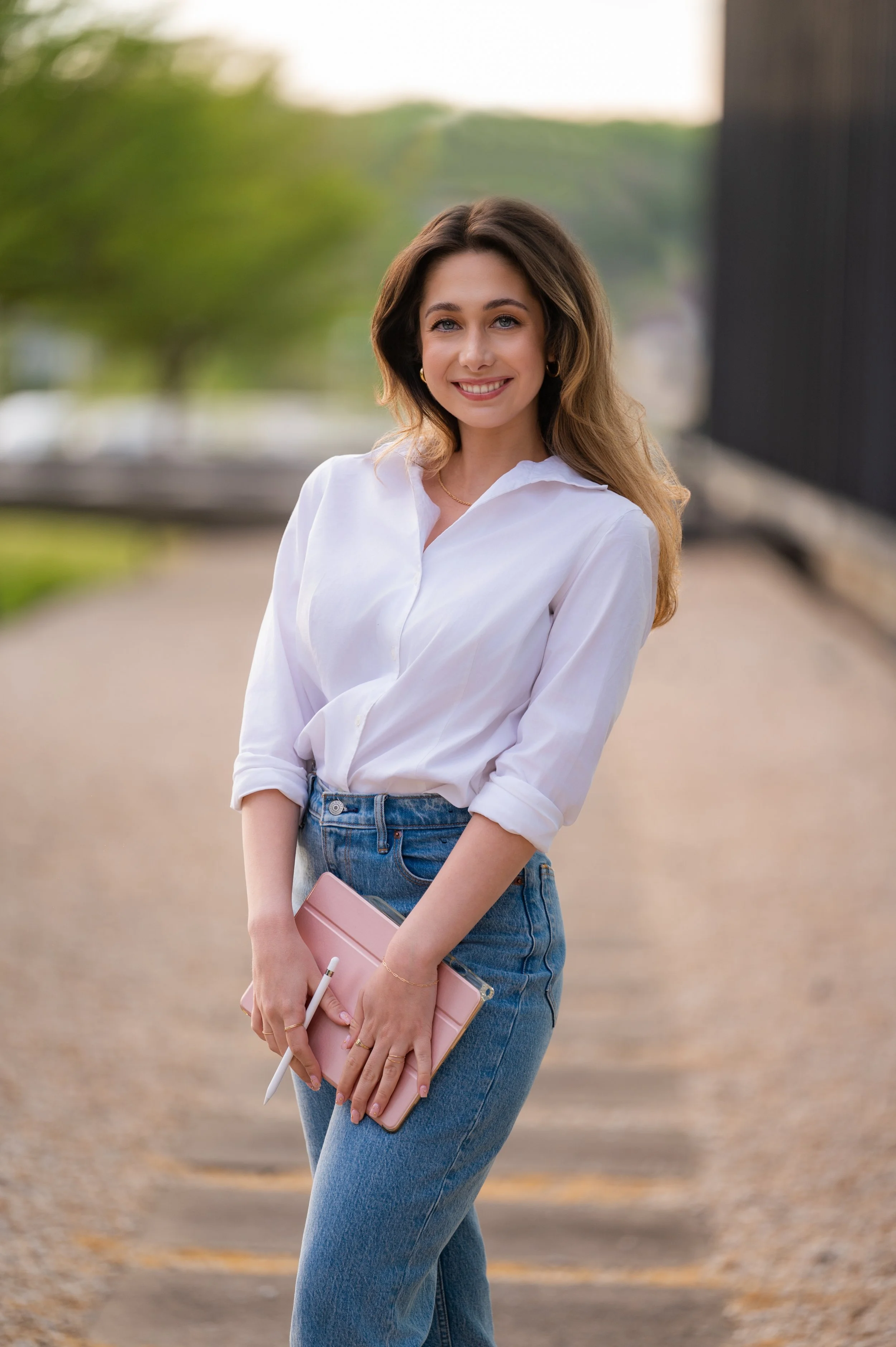 A smiling young woman with wavy brown hair, dressed in a white blouse and blue jeans, holding a pink tablet and stylus, standing outdoors on a paved walkway with trees and a black fence in the background.