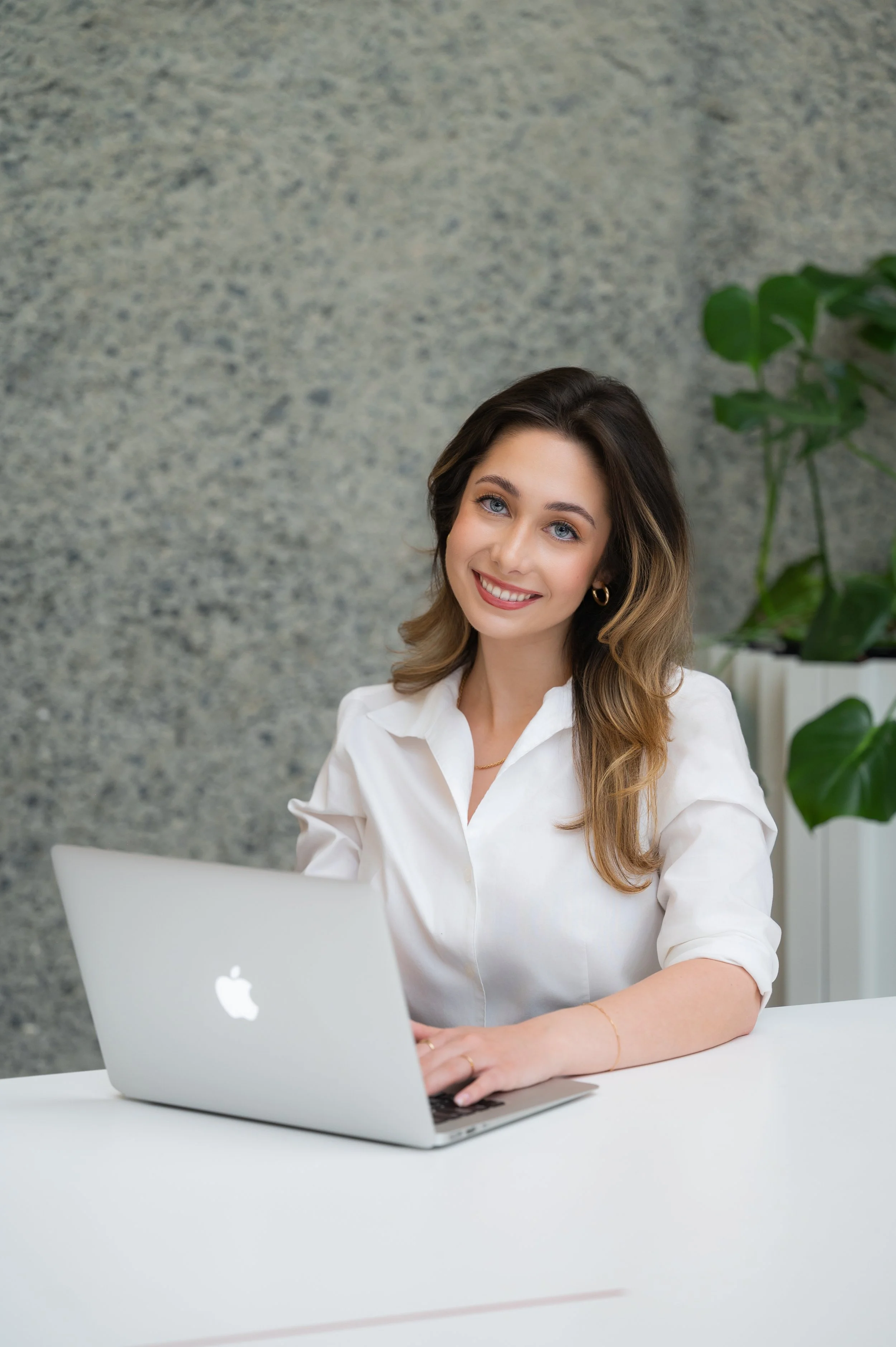 A smiling woman with shoulder-length wavy brown hair and blue eyes, wearing a white blouse, sitting at a white desk with a silver MacBook laptop, in front of a textured gray wall and green plant in the background.