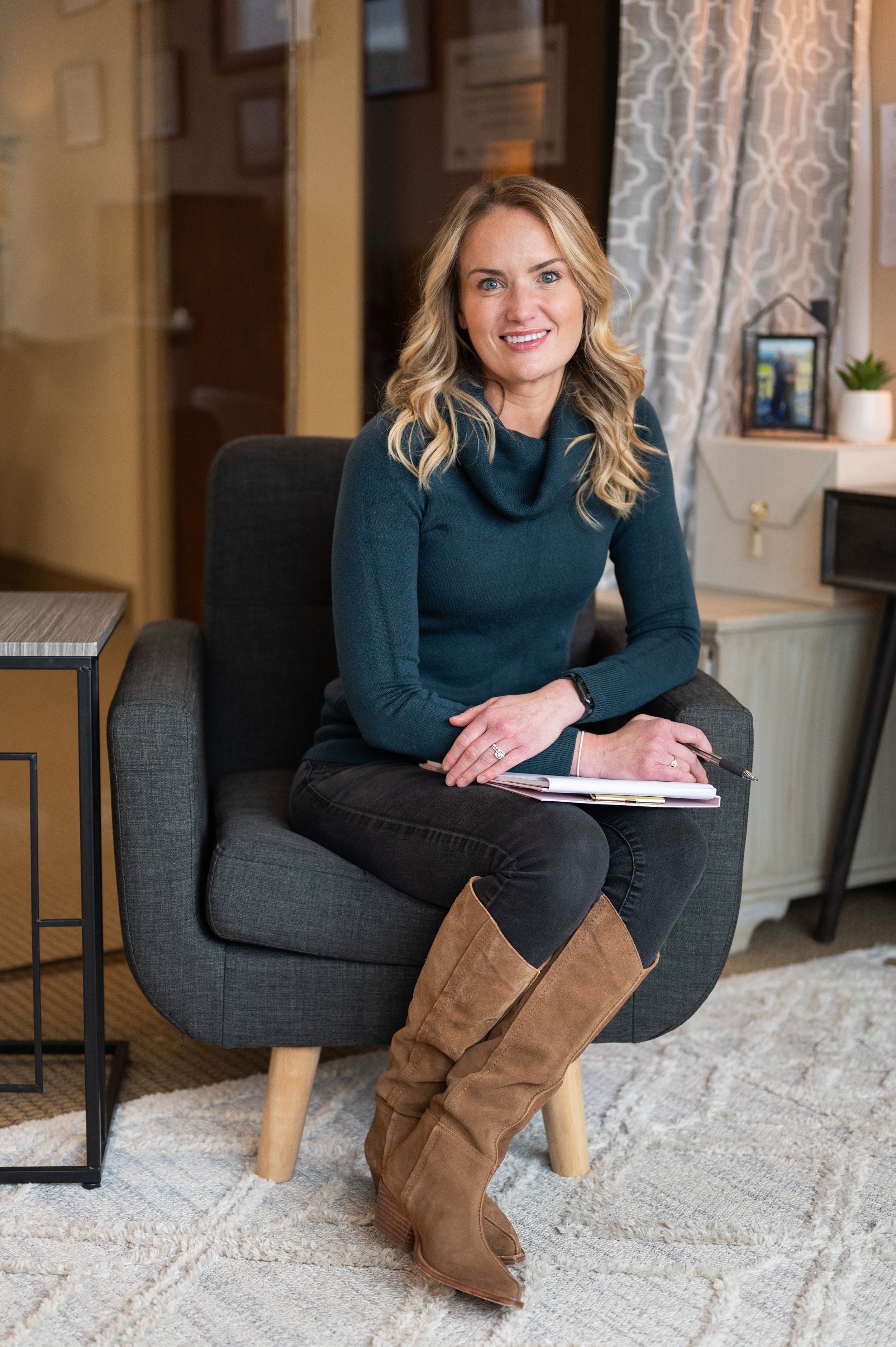 A woman with blonde wavy hair, wearing a teal turtleneck sweater, black pants, and brown boots, is sitting on a dark gray armchair in a cozy room with a patterned beige rug, a small side table, and a cabinet with framed pictures and plants.