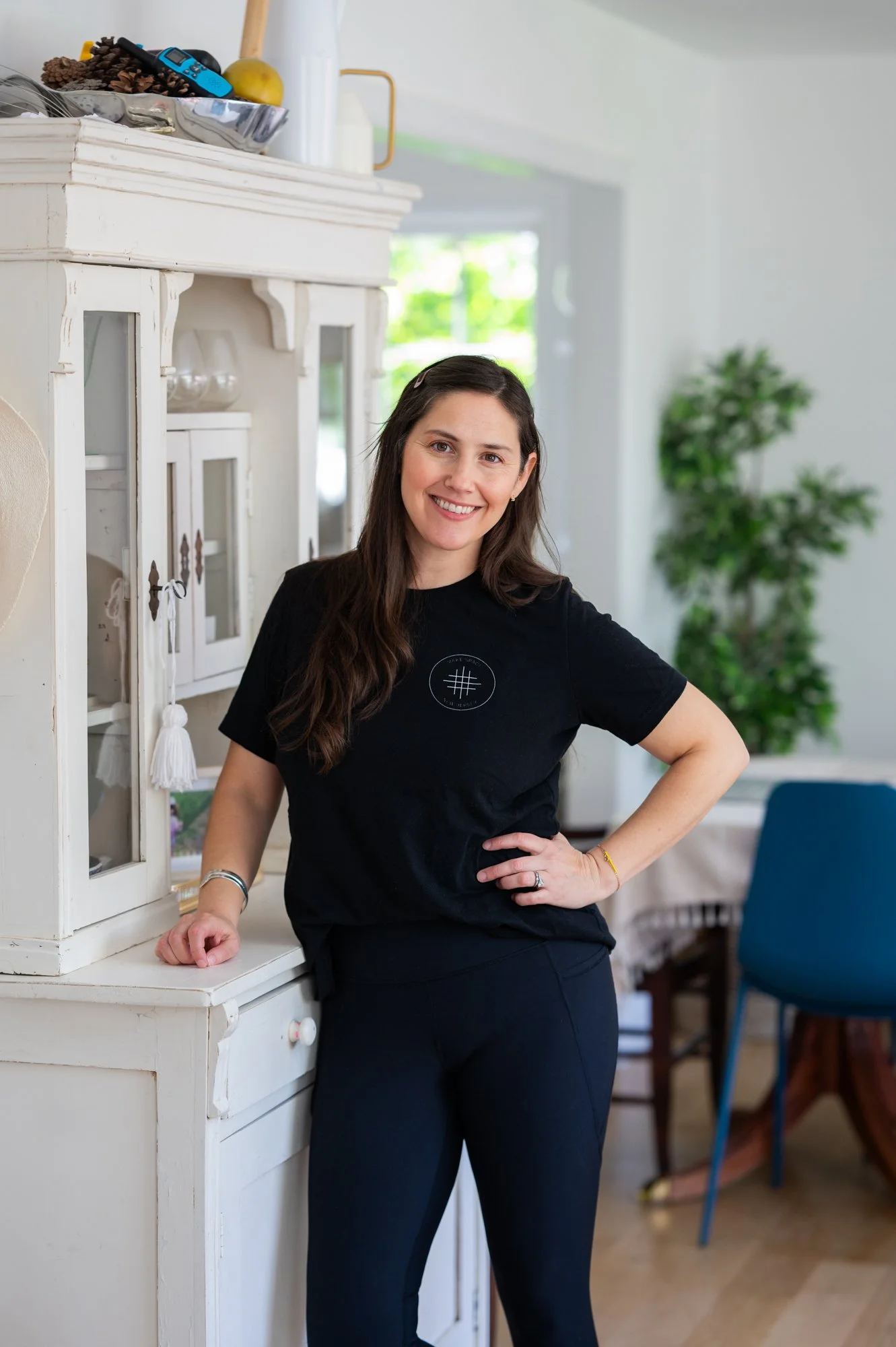 A woman with long dark hair wearing a black T-shirt and black pants standing in a brightly lit room, smiling, with a white cabinet and a blue chair in the background.