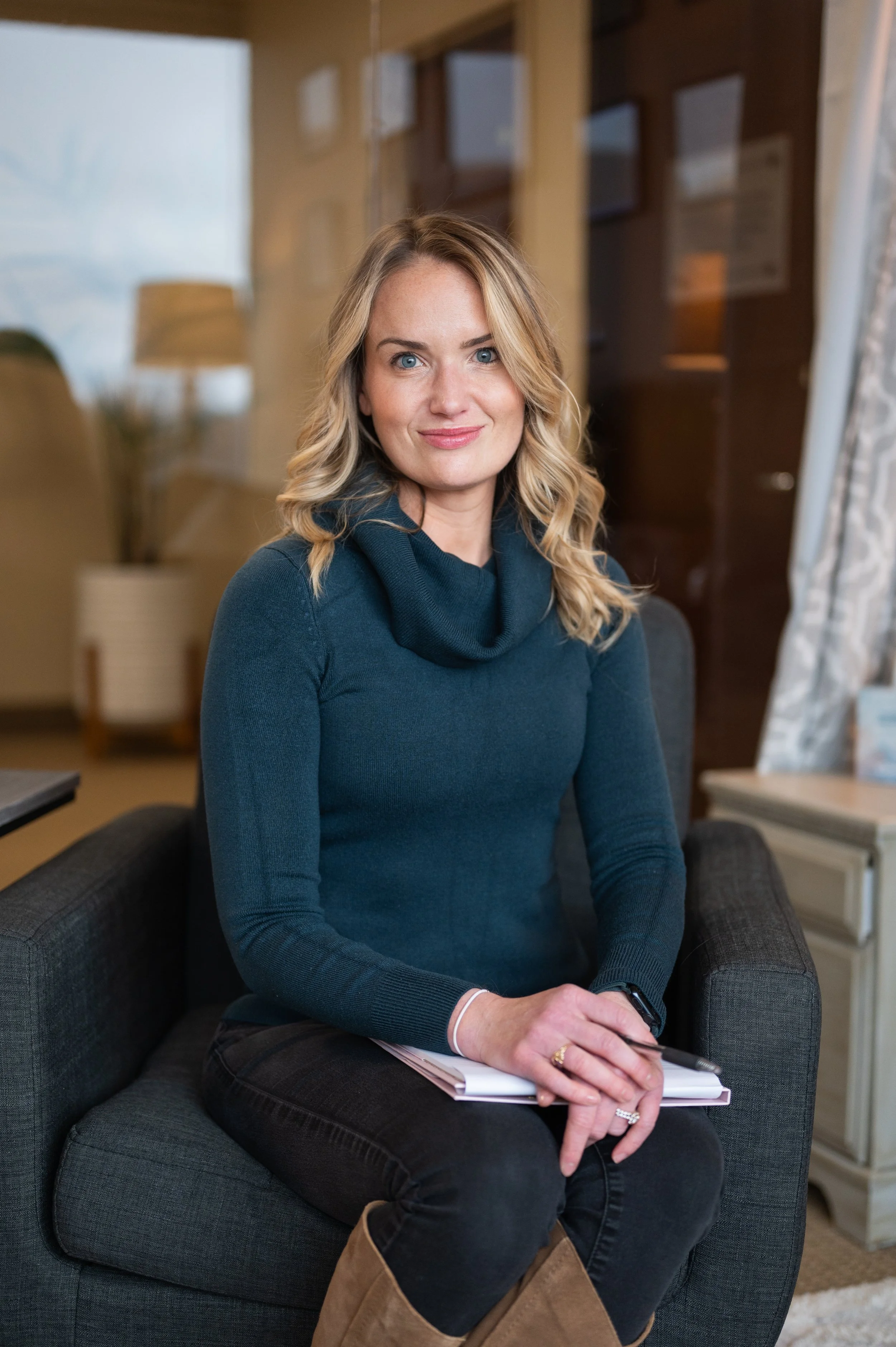 A woman with blonde hair and blue eyes sitting on a black armchair in a cozy, well-lit room. She is wearing a dark teal sweater with a cowl neck, black pants, and brown boots, holding a notebook and pen. The background shows a living room with booksh