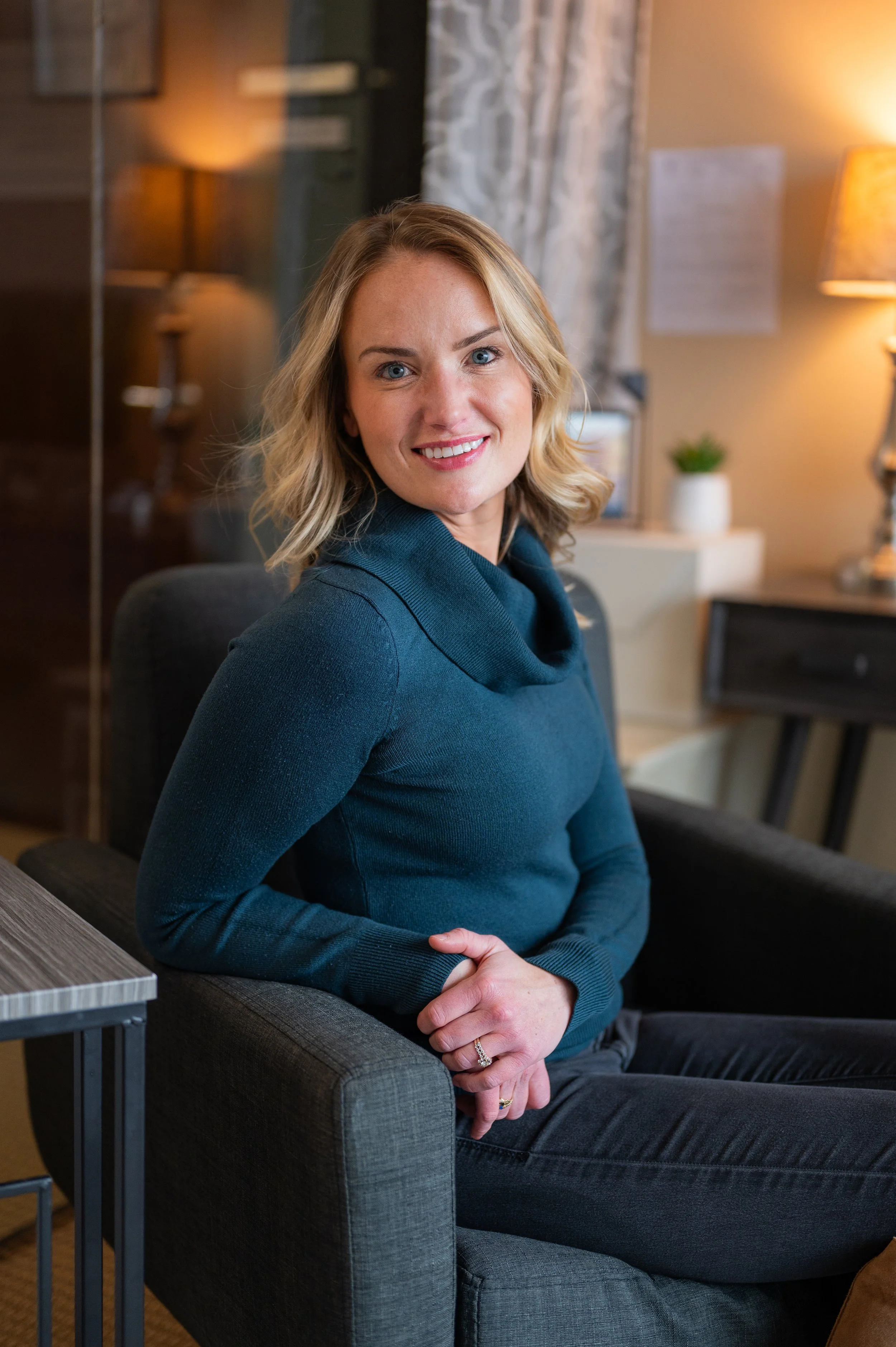 A woman with blonde hair sitting in an armchair, smiling, wearing a teal sweater in a cozy room.