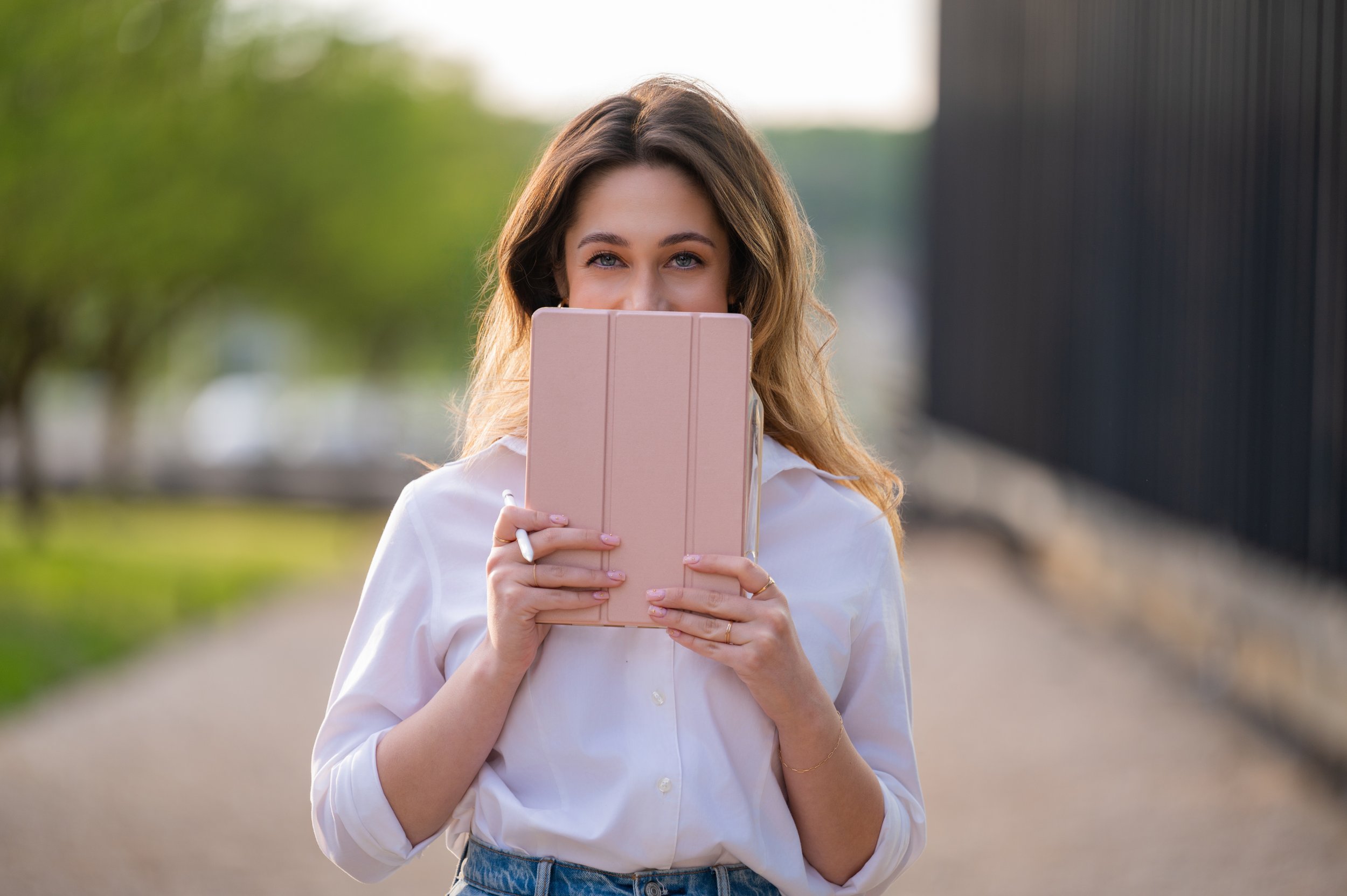 A young woman outdoors holding a pink tablet device with both hands, partially covering her face, and looking at the camera.