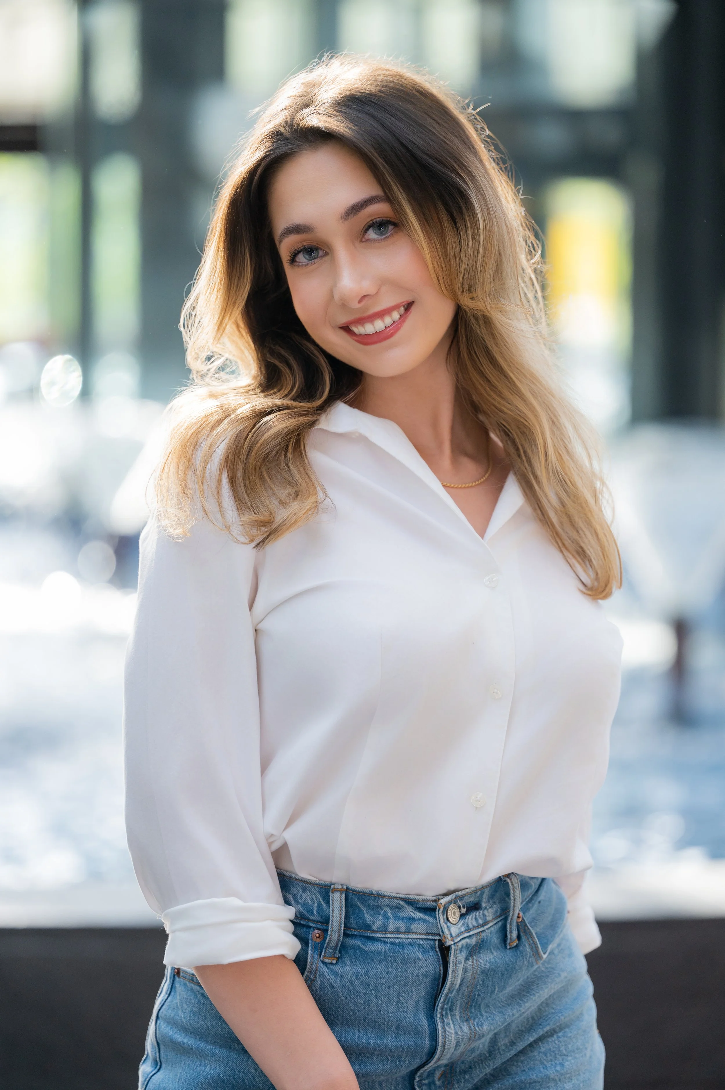A young woman with long, wavy light brown hair and blue eyes, smiling, wearing a white button-up shirt with sleeves rolled up and blue jeans, standing indoors near large windows with blurred outdoor scenery in the background.