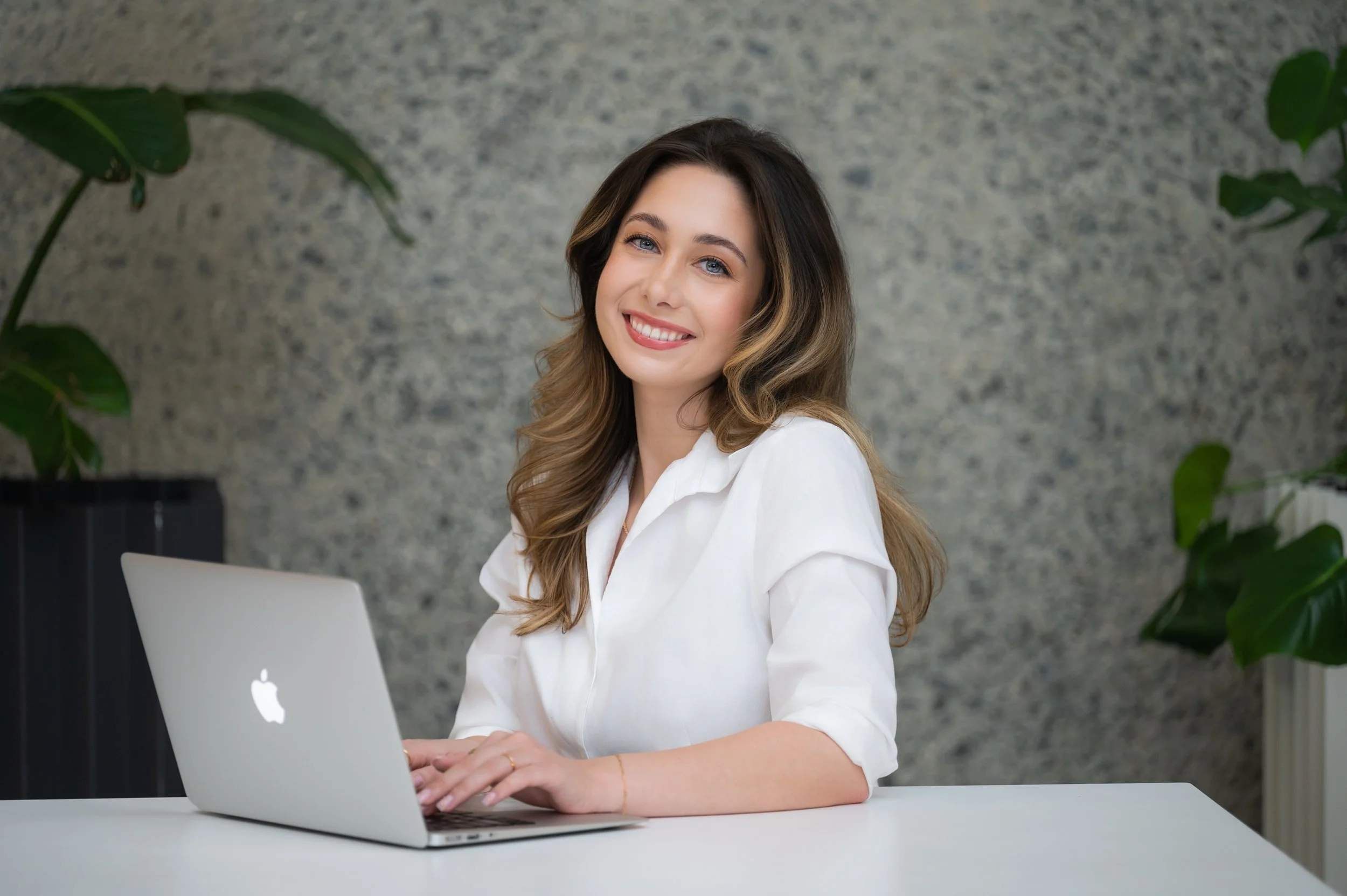 A woman with long, wavy brown hair sitting at a white table using a silver laptop with a white Apple logo. She is smiling and wearing a white shirt, with a textured gray wall and green plants in the background.