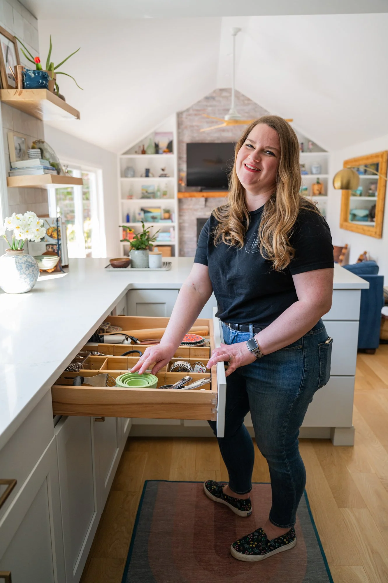 A woman standing in a kitchen, opening a drawer with kitchen utensils, smiling at the camera.