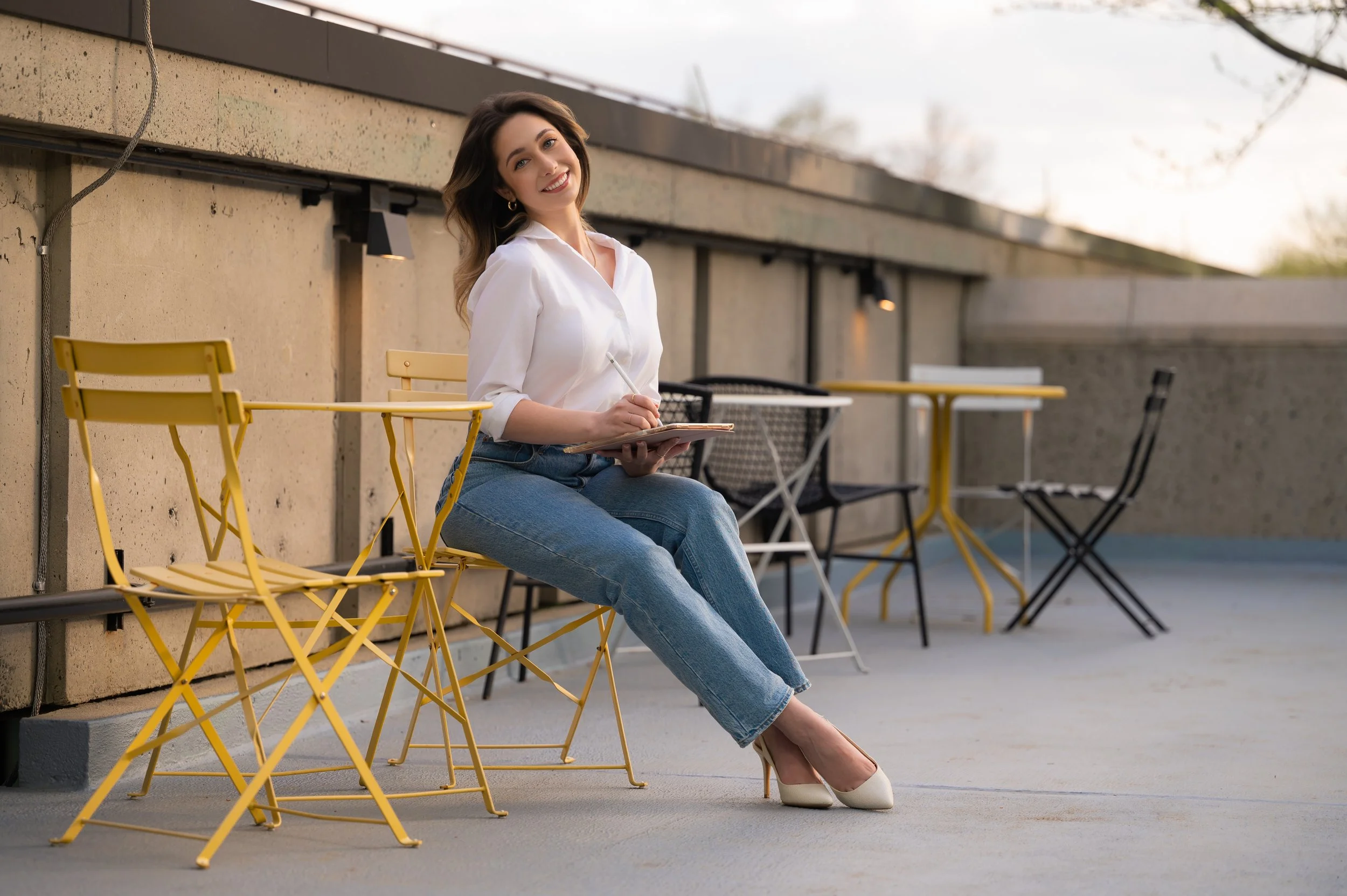 A young woman sitting on a yellow outdoor chair on a rooftop terrace, smiling at the camera, holding a notepad and pen, wearing a white shirt, blue jeans, and white heels, with additional yellow and black chairs and tables around her.