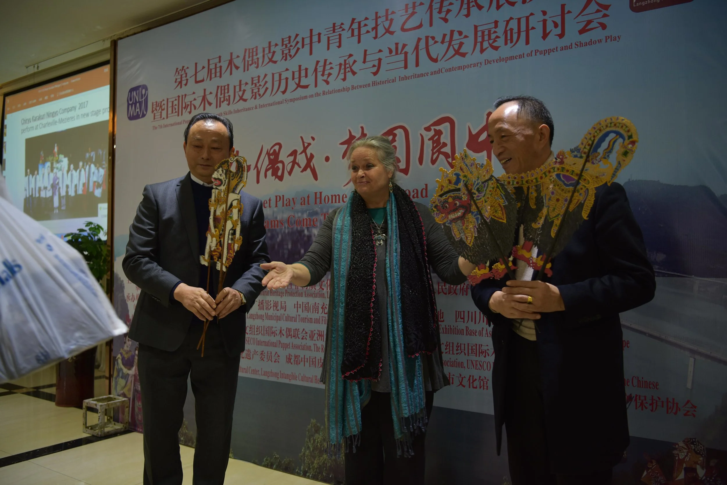 Photo courtesy of UNIMA CHINA. (Left to Right): After introducing the 2020 Bali Congress and World Puppetry Festival, Karen Smith presents two Balinese shadow puppets (Hanuman and Barong) to Wang Biao (Director, Sichuan Wang’s Shadow Puppet Troupe) and Wang Fang (Sichuan Wang’s Shadow Puppet Troupe).