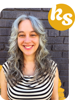 A photo of me smiling at you. I'm a white woman with long wavy salt & pepper hair wearing a black and white striped shirt, standing in front of a gray brick wall.