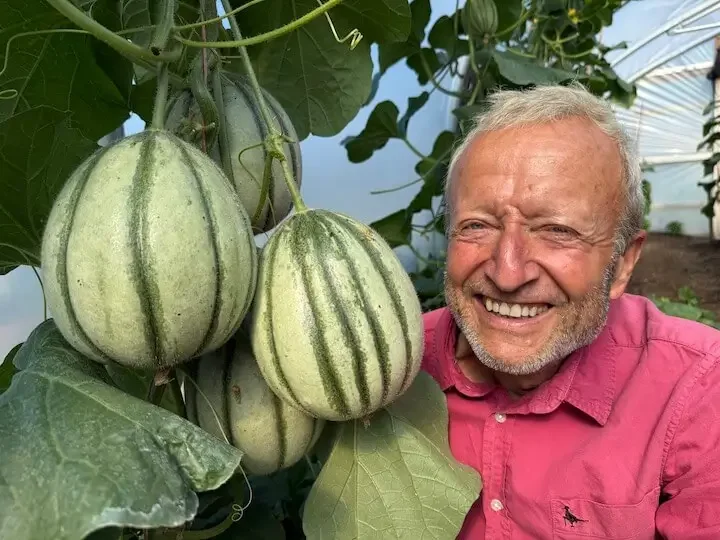 Charles Dowding smiles beside a vibrant display of squash in a poly tunnel garden.