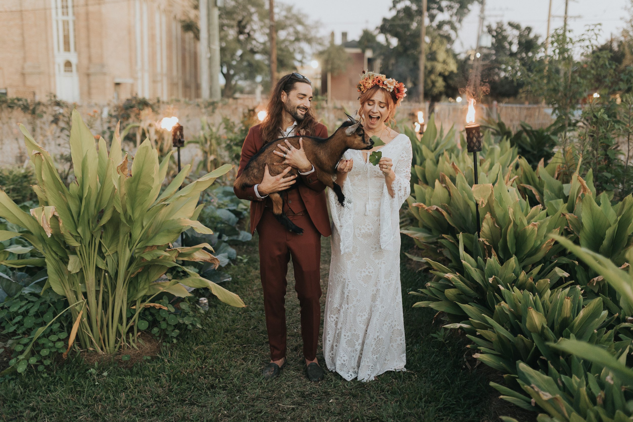 A bride and groom posing with a Pygmy goat, with the backdrop of the lush greenery of Paradigm Gardens, a garden wedding venue in New Orleans.