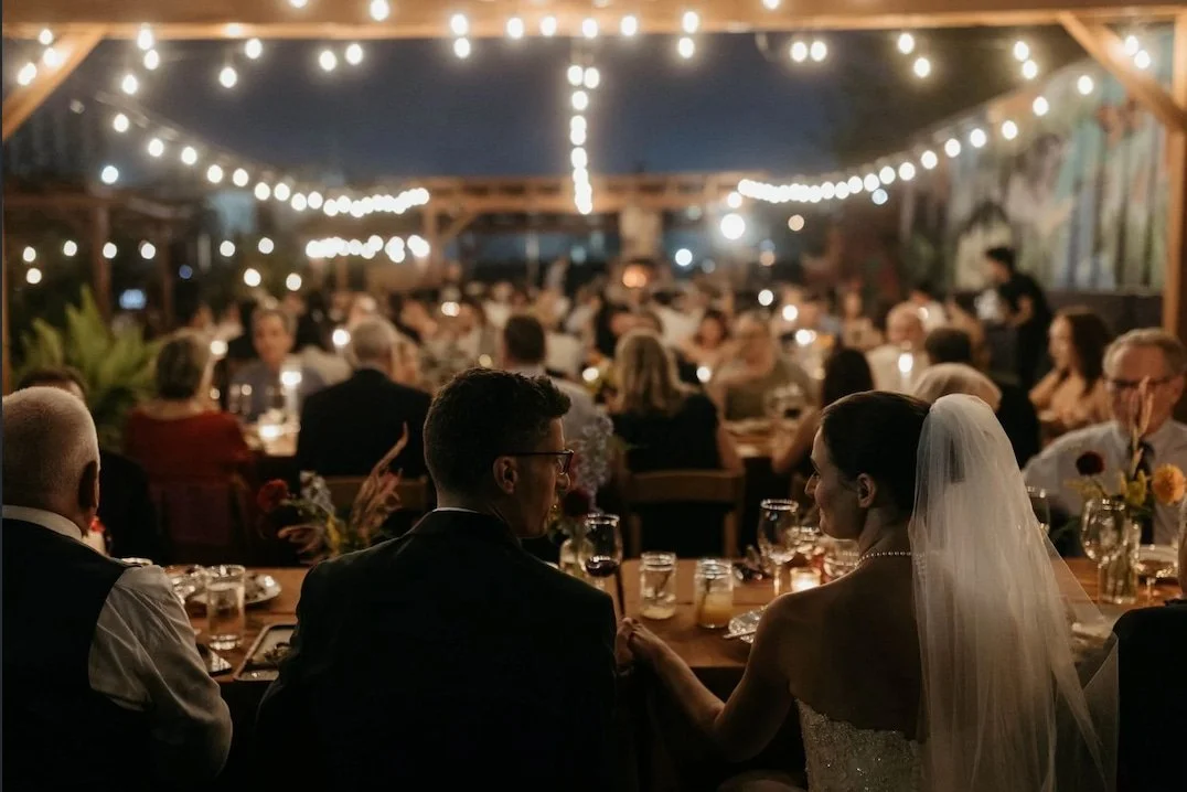 Bride and groom sitting after wedding ceremony overlooking guests in outdoor venue under string lights.