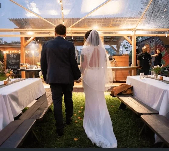Bride and groom holding hands under clear tent in outdoor garden venue.