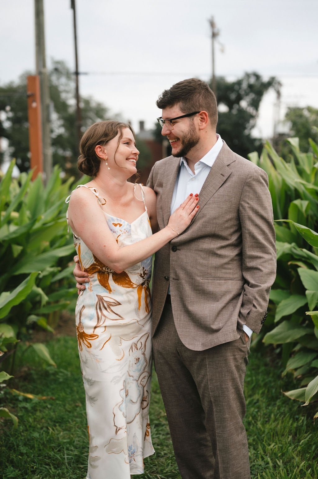 A smiling couple standing together outdoors, with green plants in the background. The woman is wearing a floral dress, and the man is in a brown suit.