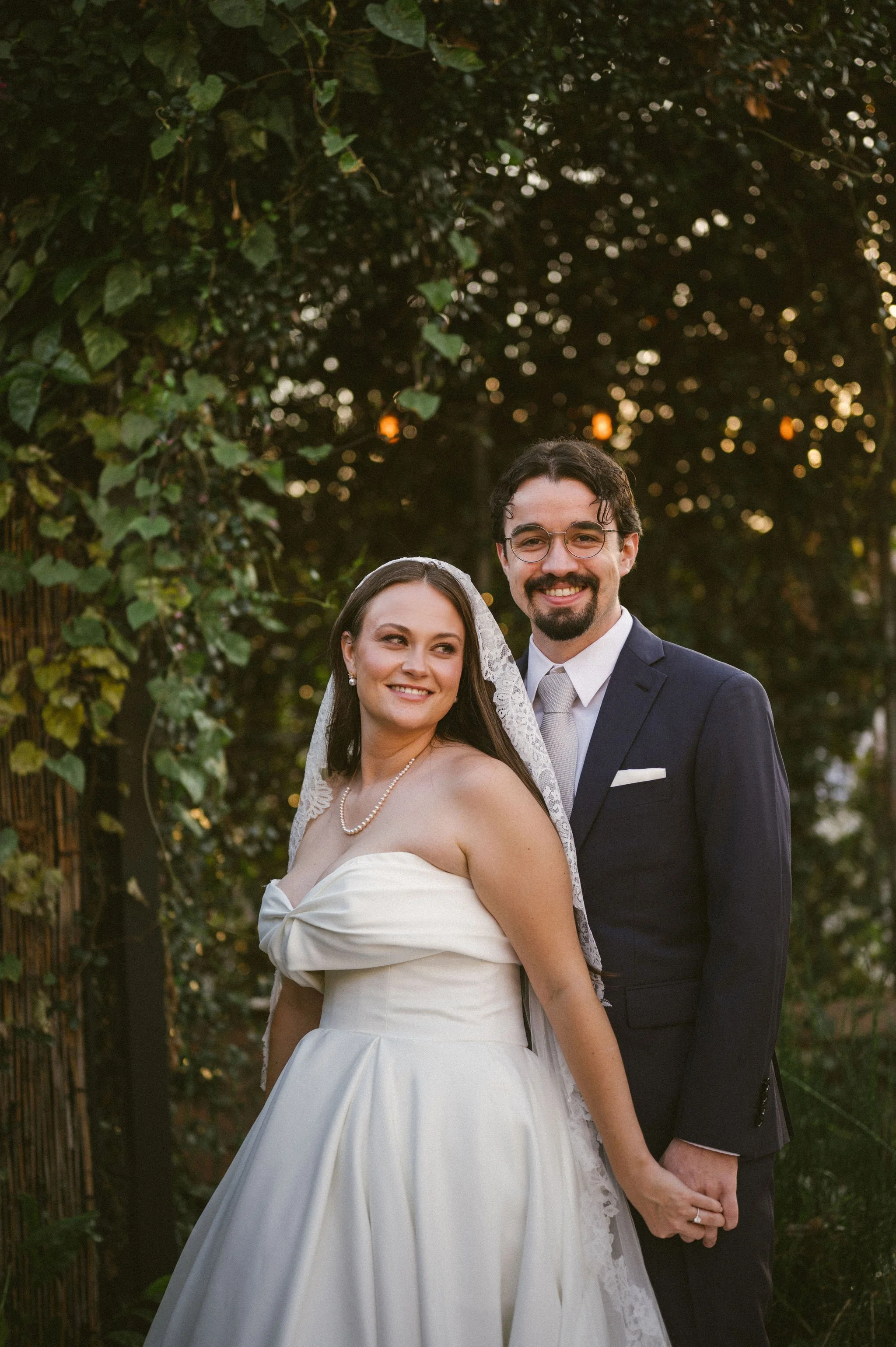 A bride and groom smiling and holding hands outdoors during sunset, with greenery and string lights in the background.