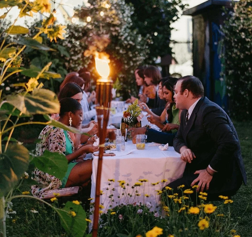 Group of wedding guests sitting down and eating farm-to-table dinner in outdoor garden venue.