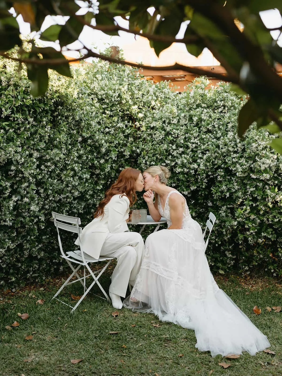 Two brides sitting down kissing in front of jasmine flower wall.