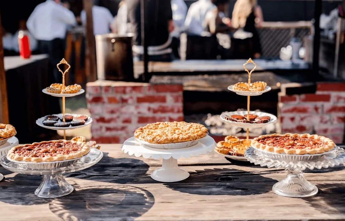 Mixed pies and other desserts spread out on wood table with wood-fire grill in the background.
