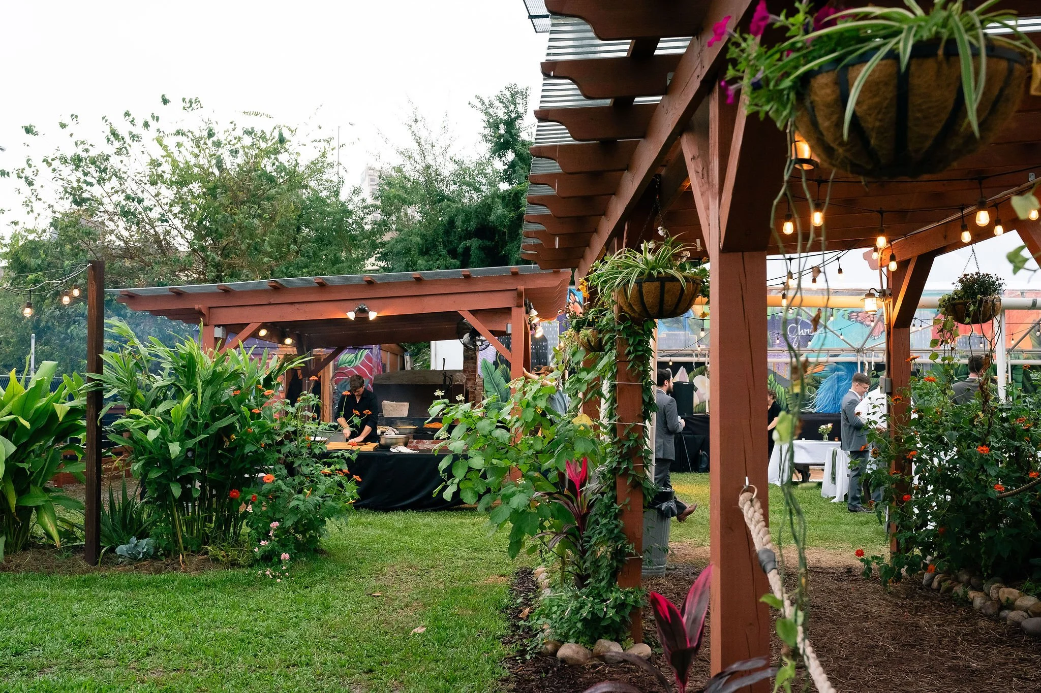 Outdoor garden event with wooden pergolas, hanging plants, string lights, and people socializing. A chef is preparing food in the background.