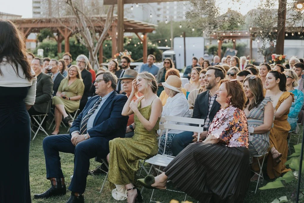 Outdoor wedding and gathering with people seated, smiling and attentive, under string lights during daytime.